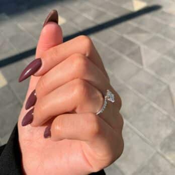 תקריב של יד המציגה את דגם טבעת אירוסין הנוצץ לוליטה ב-40% הנחה, עם ציפורניים מטופחות צבועות בגוון סגול כהה, מתמוגגות.