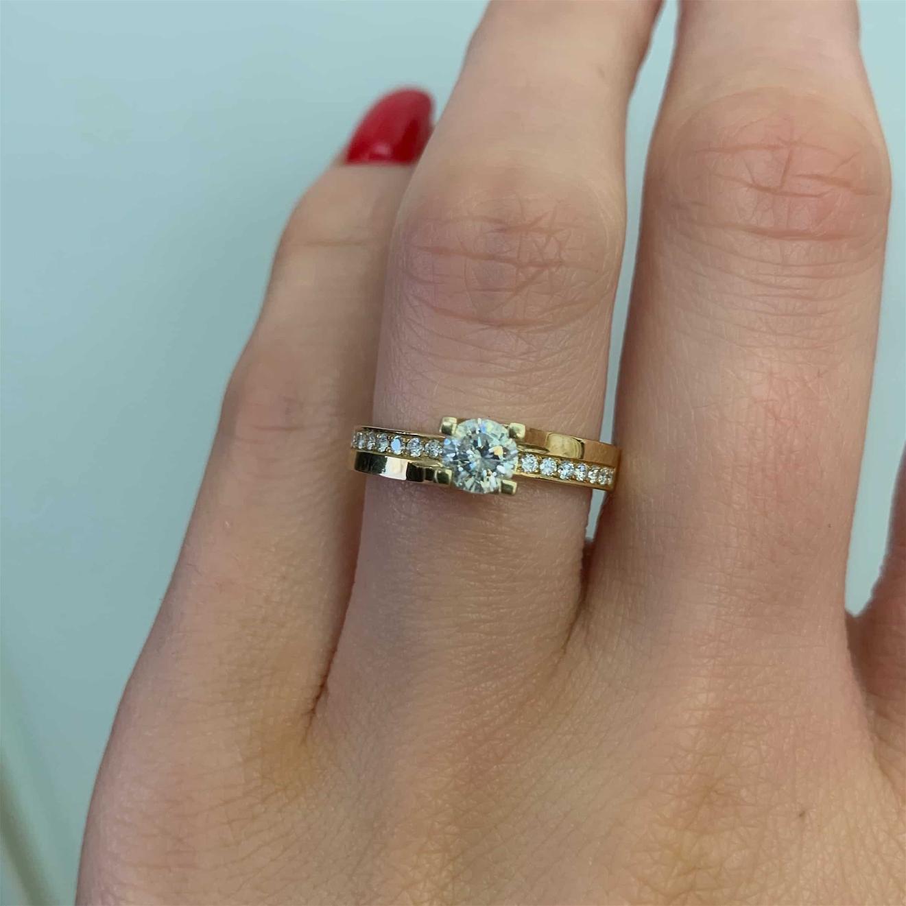 תקריב של יד המציגה את הזהב המבריק טבעת אירוסין דגם ימית ב-38% הנחה טבעת אירוסין עם יהלום מרכזי בולט ויהלומים קטנים נוספים משובצים בלהקה.
