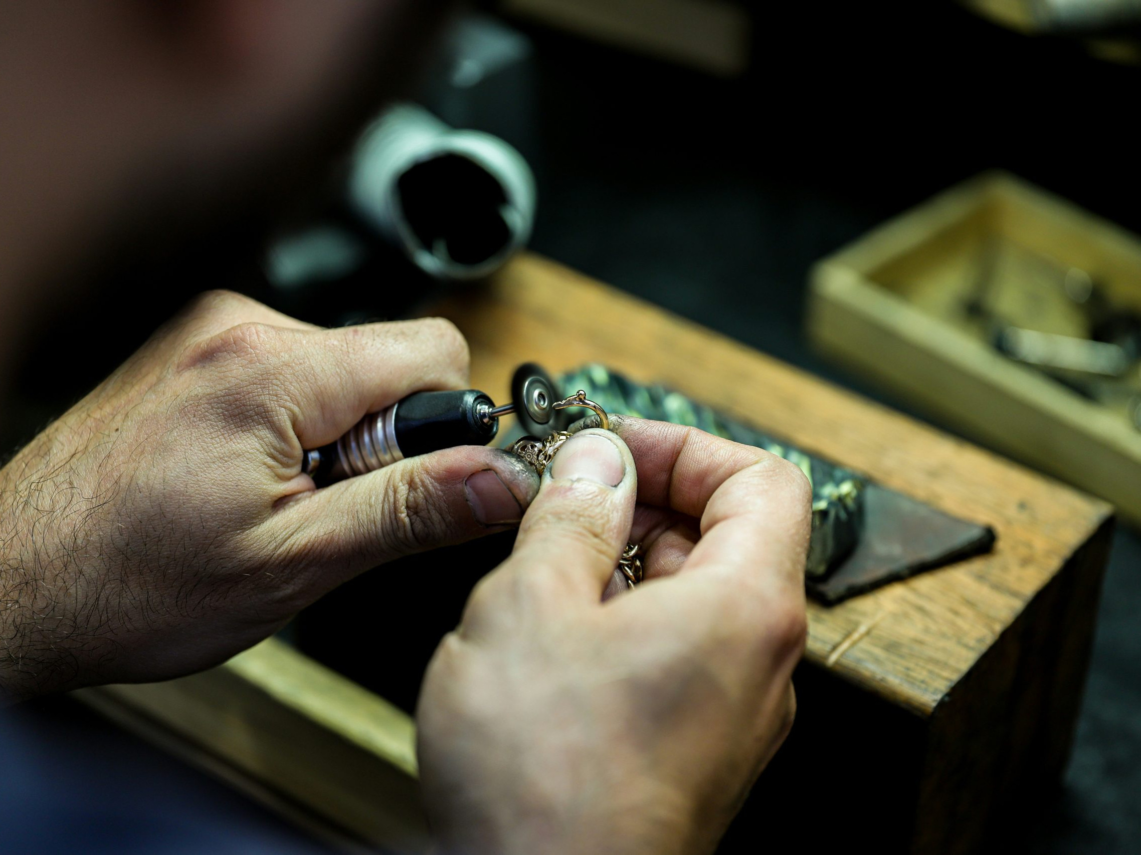 תקריב של ידיו של גמולוג עובד בקפדנות על תיקון תכשיט קטן ומורכב באמצעות כלי עבודה, על שולחן עבודה עמוס.